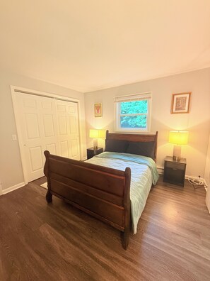 Quarto com cama de madeira, duas mesas de cabeceira com luminárias, janela com vista para o verde e uma porta.