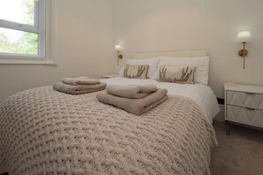 A neatly made bed with a knitted blanket and two folded towels, a bedside table with a lamp, and a window with a view of trees. - Boutique Apartment Little Gem Hessle, ground floor modern fully renovated 2026 (Hessle)