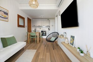 Een moderne woonkamer met een bank, een eettafel met stoelen, een zwarte geweven stoel, een keuken met kasten, en een aan de muur bevestigde televisie.