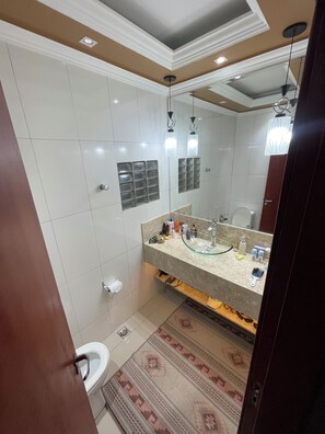 Un baño con un lavabo de vidrio, azulejos blancos, un inodoro y una alfombra estampada.