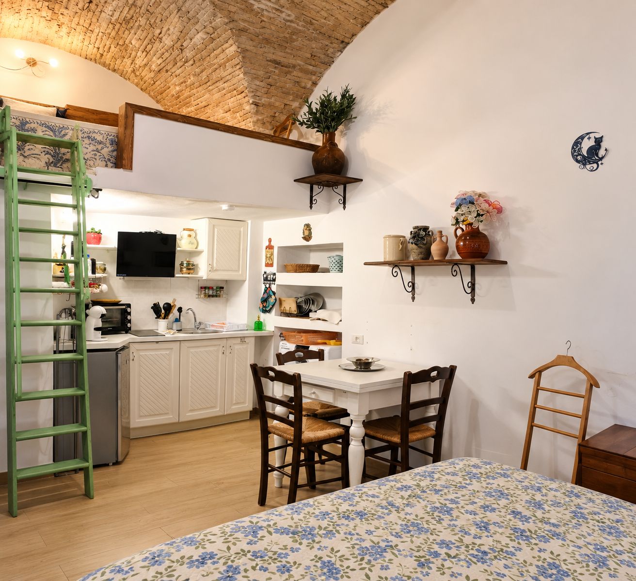 Een compacte keuken met witte kasten, een eettafel met stoelen, een groene ladder en een aan de muur bevestigde televisie.