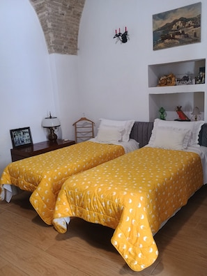 A room with two beds, a wooden floor, a brick wall, a painting, and a shelf with various items.