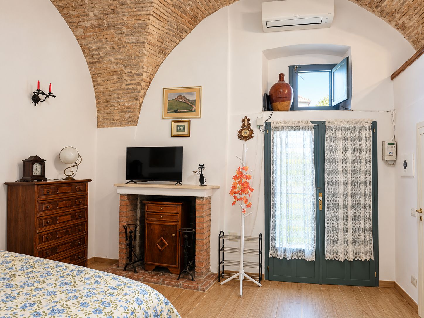 Een kamer met een bakstenen boog, een televisie op een schoorsteenmantel, een bed met een bloemenbedekking, een houten dressoir en een deur met gordijnen.