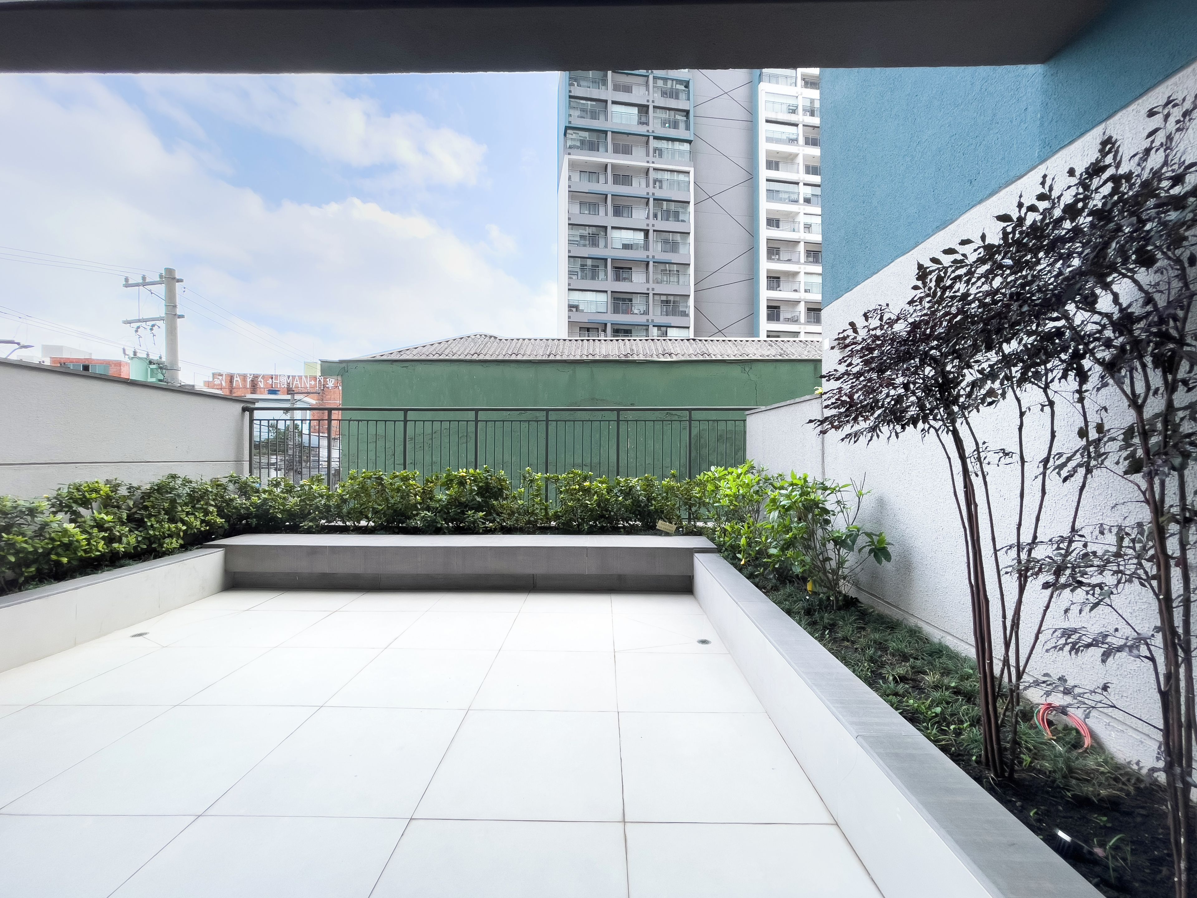 A rooftop terrace with a view of surrounding buildings, a green fence, and some vegetation.