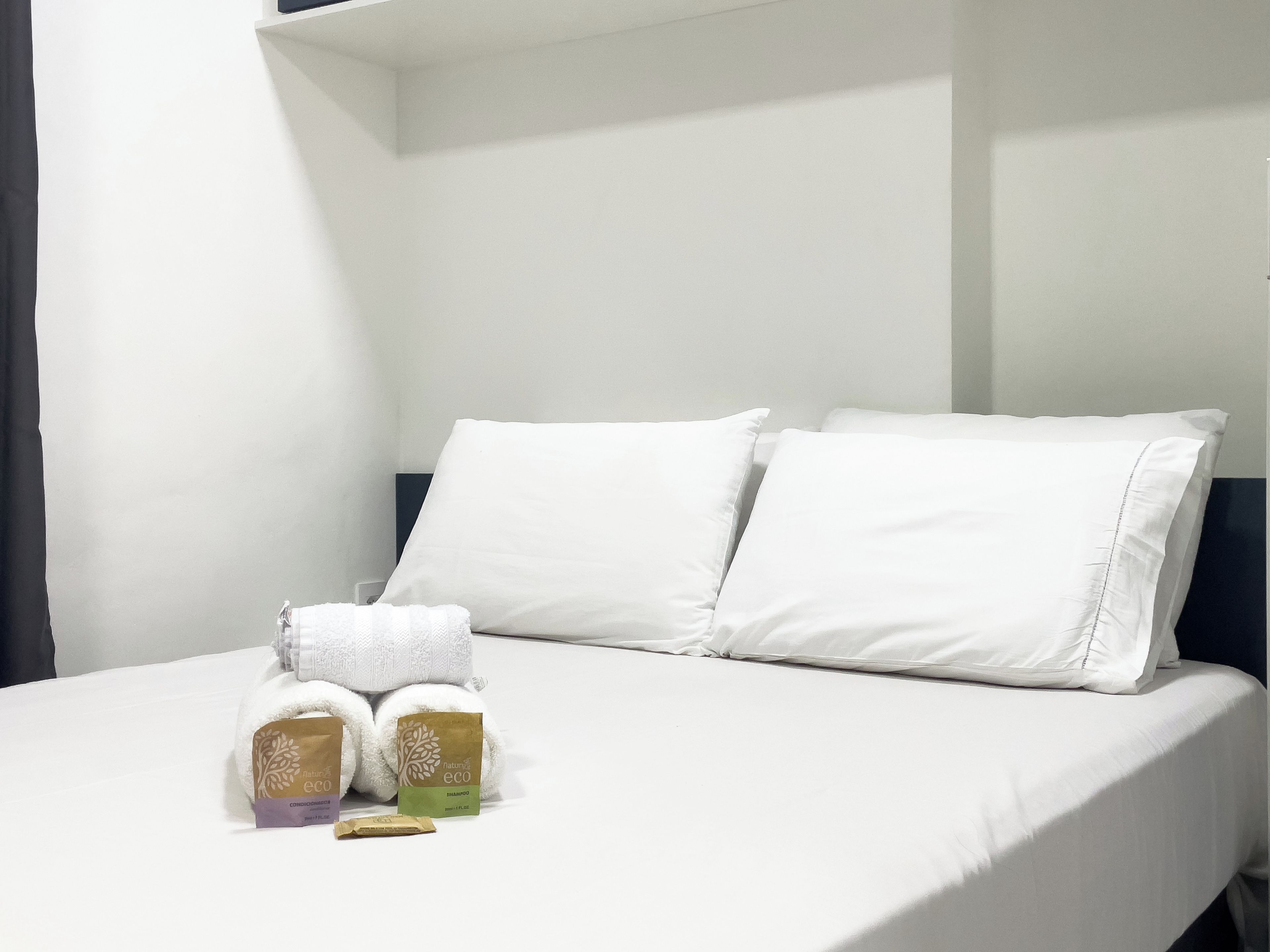 A neatly made bed with white linens and two pillows, a set of towels, and two small bags of shampoo and conditioner.