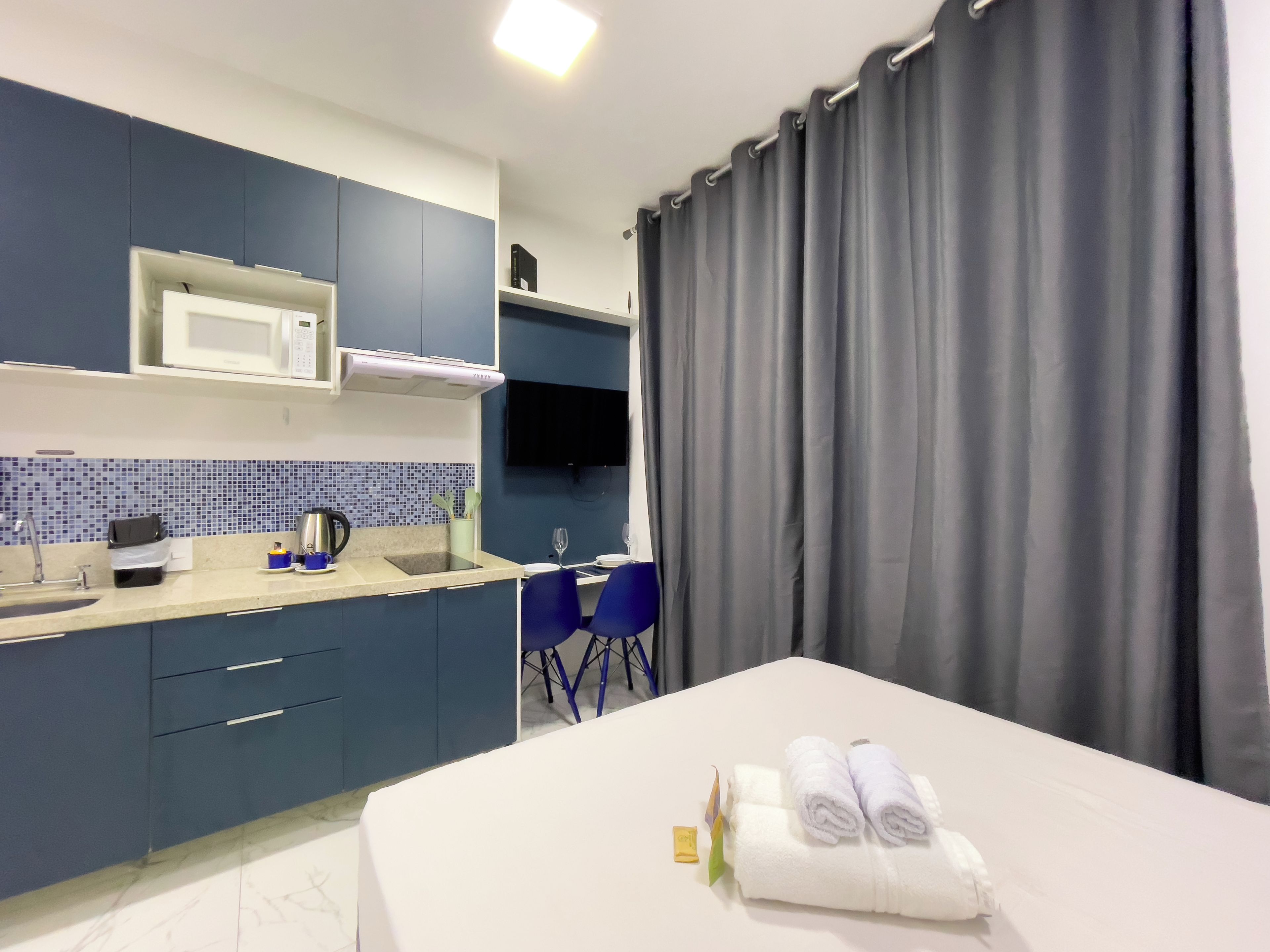 A modern hotel room with a bed, a small kitchenette, a flat-screen TV, and a window with curtains.