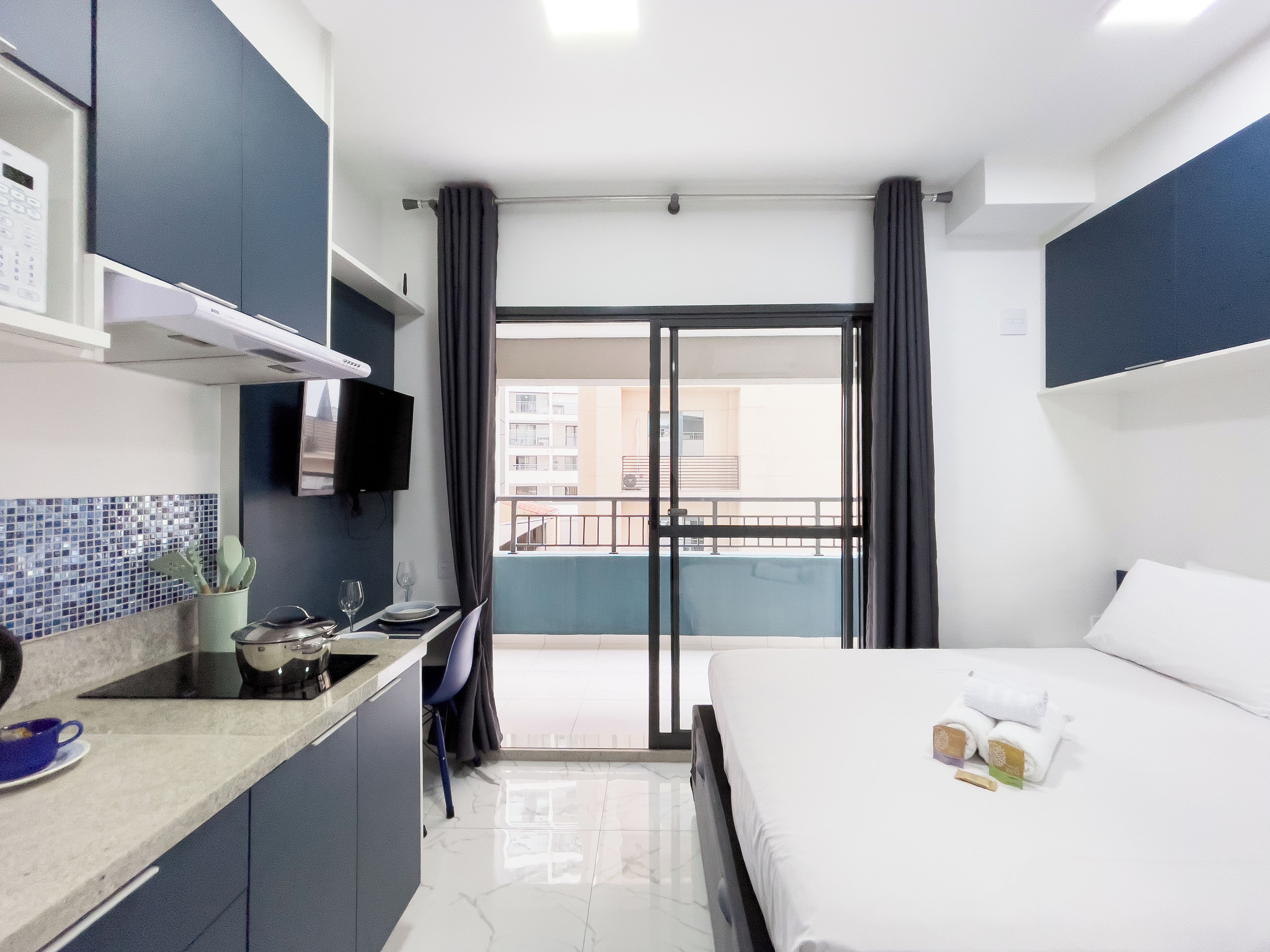 A hotel room with a bed, a small kitchenette, a balcony with curtains, and a view of buildings.