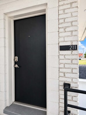 A black door with a numeric address sign '211' on a white brick wall. - Sandy Serenity at Tenth | Unit 211 (Myrtle Beach)