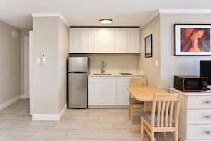 A compact kitchen with a refrigerator, sink, and microwave. There is a dining table with chairs and a framed picture on the wall. - Sandy Serenity at Tenth | Unit 211 (Myrtle Beach)