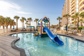 A pool area with a children's play structure, including a slide, surrounded by palm trees and lounge chairs. - Ocean Walk Resort 2023 A & B (Daytona Beach)