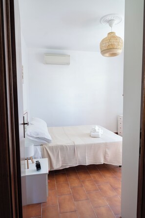 Una habitación pequeña y limpia con una cama individual, una mesita de noche, aire acondicionado de pared y una lámpara de techo tejida.