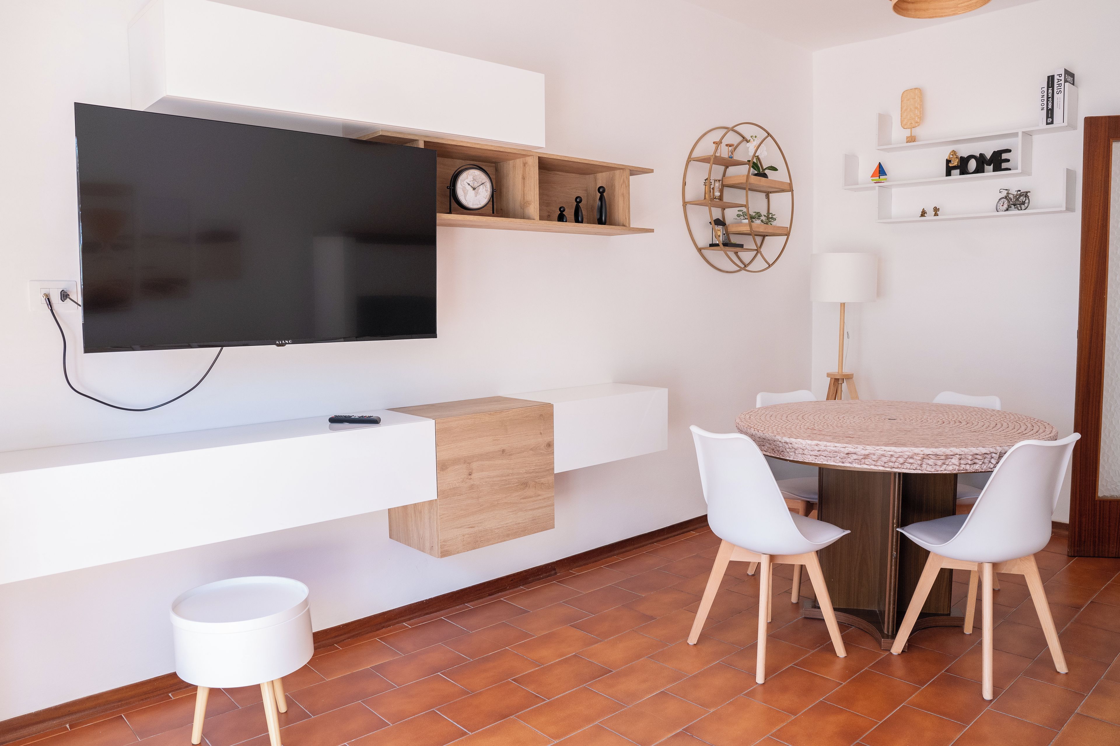 Un espace de vie moderne avec un téléviseur à écran plat fixé à un mur blanc, une étagère en bois ornée d’objets décoratifs, une table à manger ronde avec des chaises blanches et un petit tabouret blanc.