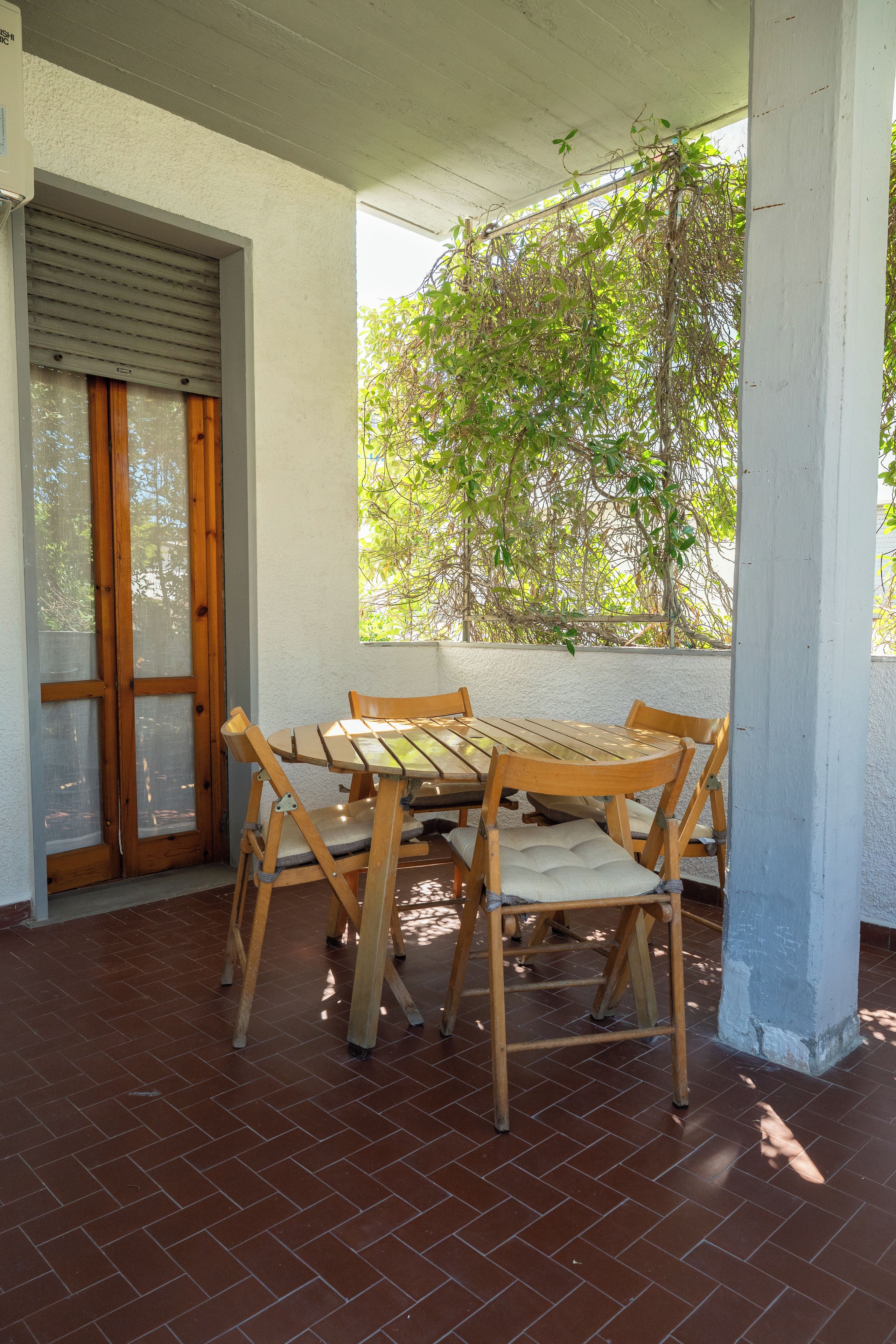 Un patio couvert avec une table et des chaises en bois, une porte-fenêtre coulissante et une vue sur la verdure environnante.