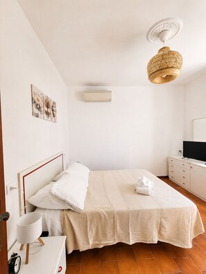 A bedroom with a bed, bedside table, lamp, air conditioning unit, artwork, and a television on a dresser.