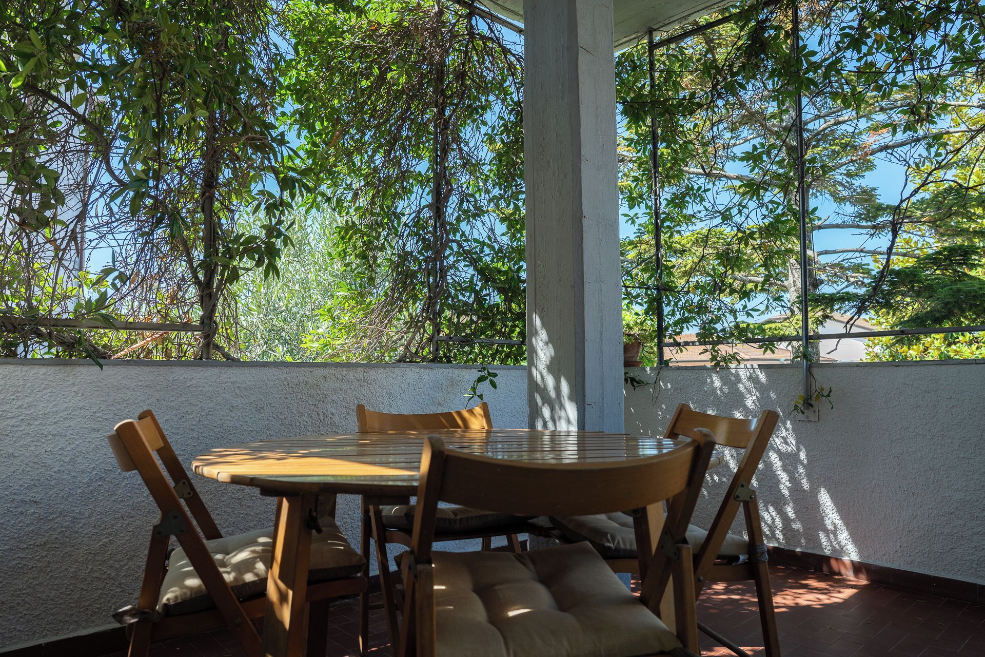 Un patio esterno con un tavolo rotondo e sedie, circondato da un muro bianco e alberi.