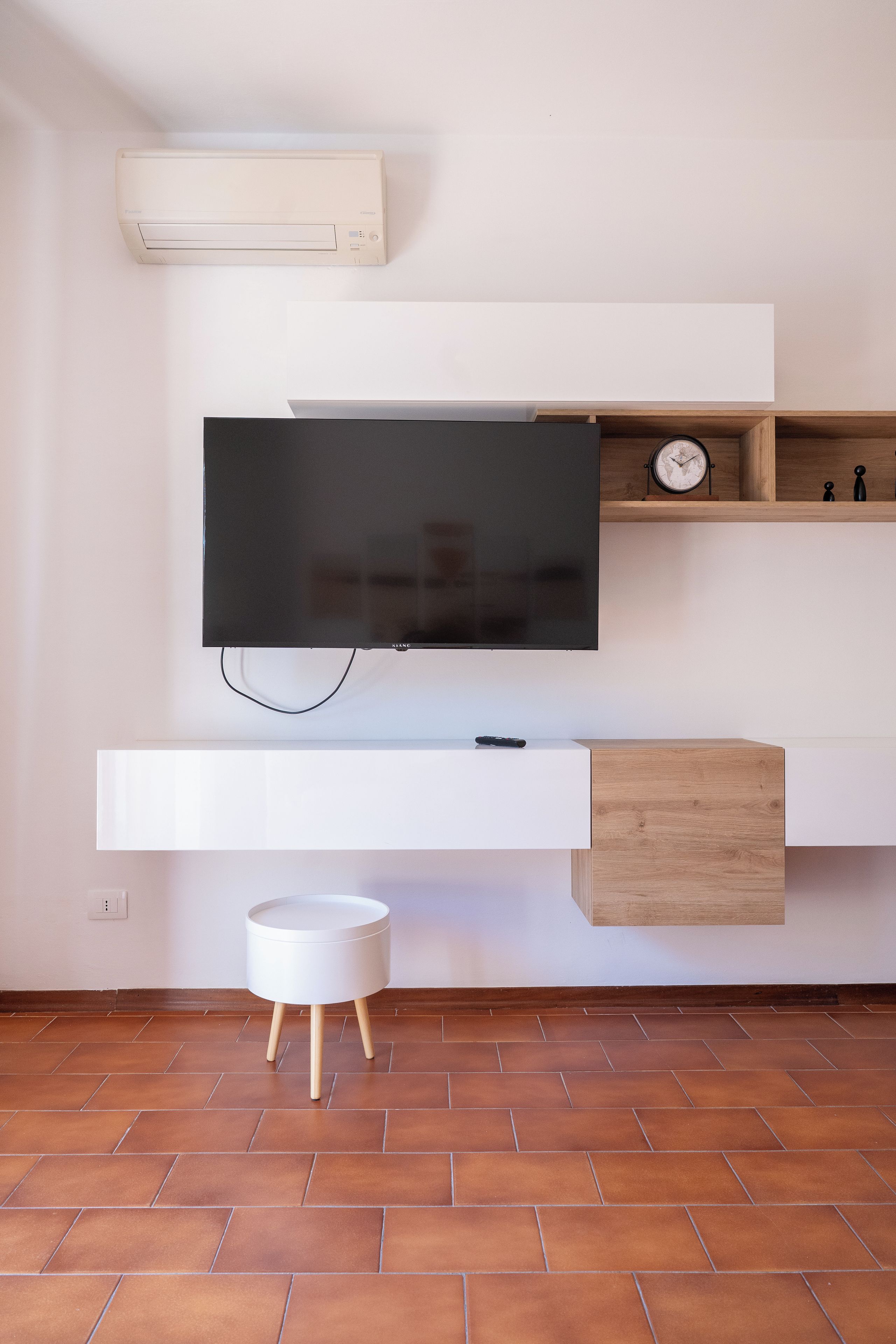 Una sala moderna con un televisor de pantalla plana montado en una pared blanca, un aparador de madera debajo y una pequeña mesa redonda blanca con patas de madera.
