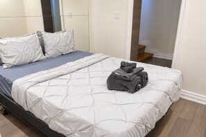 Un lit bien fait, avec une couette blanche et des oreillers gris, une serviette grise pliée et une table de chevet en bois.