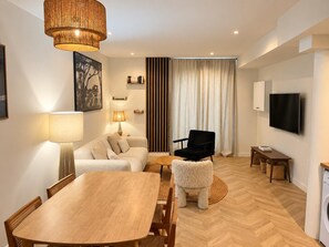 A modern living room with a wooden dining table, a sofa, a black armchair, a television, and a large window with curtains. - Central Paris – A modern and elegant flat (Paris)