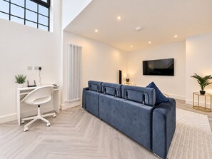 Ein modernes Wohnzimmer mit einem großen, blauen Sofa, einem weißen Schreibtisch mit Computer, einer Heizung und einem Fernseher an der Wand.