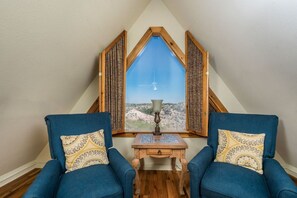 A room with two blue armchairs, a small wooden table with a lamp, and a large window with a view of a mountainous landscape.