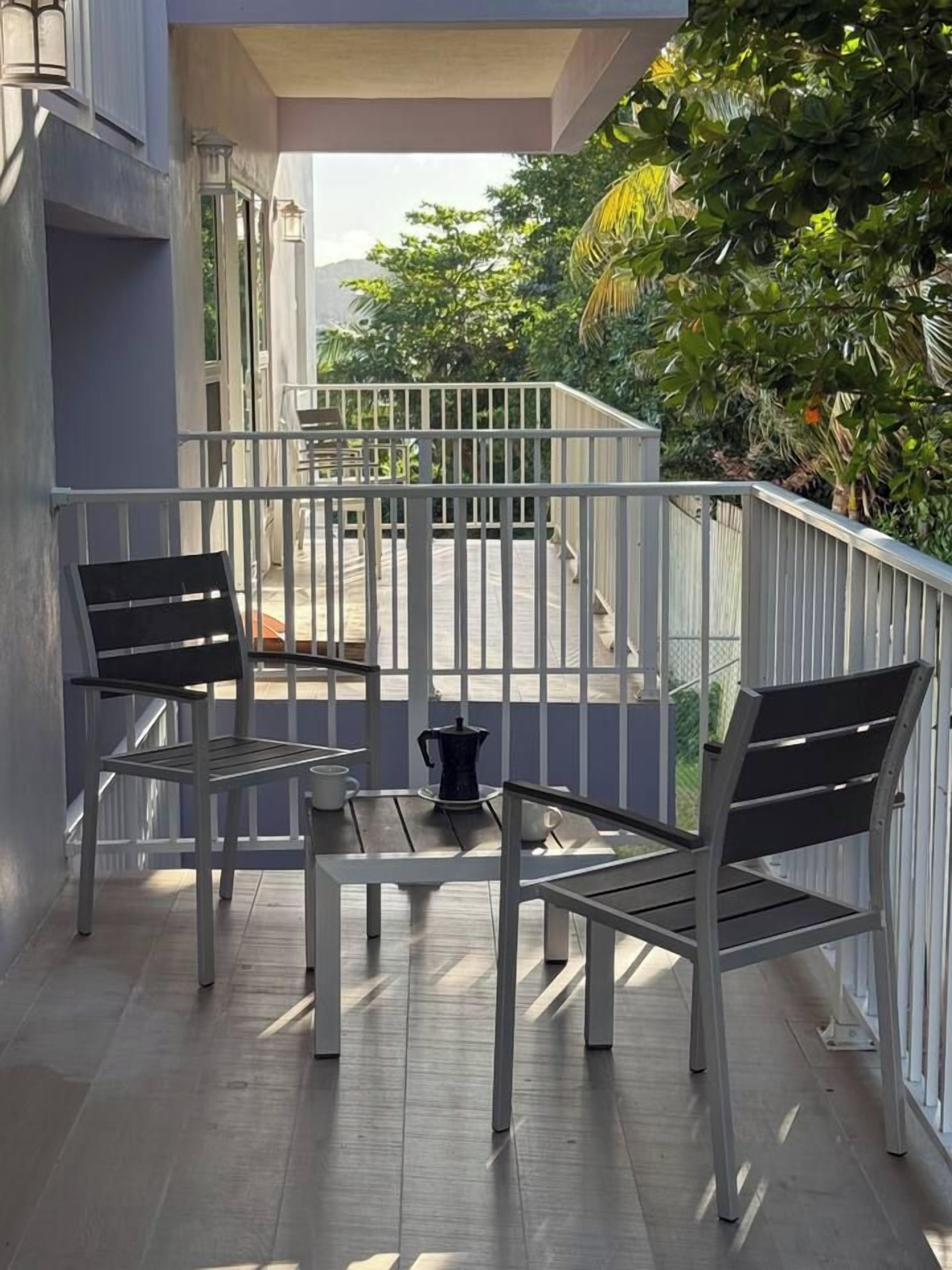 A balcony with two chairs and a table, a coffee maker, and a view of greenery and a building in the background.