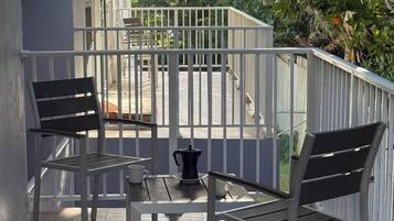 A balcony with two chairs and a table, a coffee maker, and a view of greenery and a building in the background.