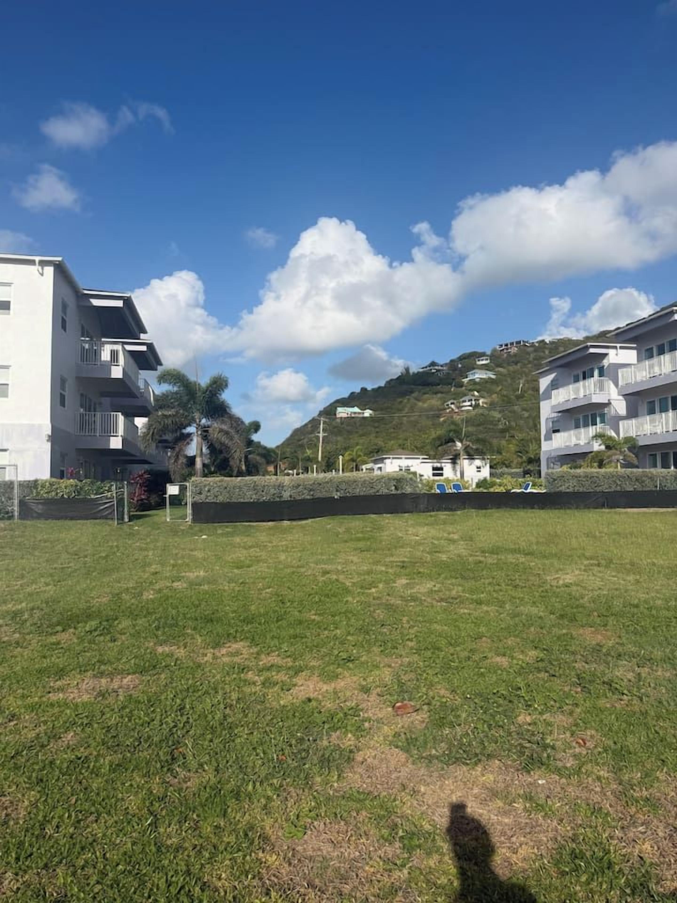 A grassy area with a building on the left and a hill with houses in the background.
