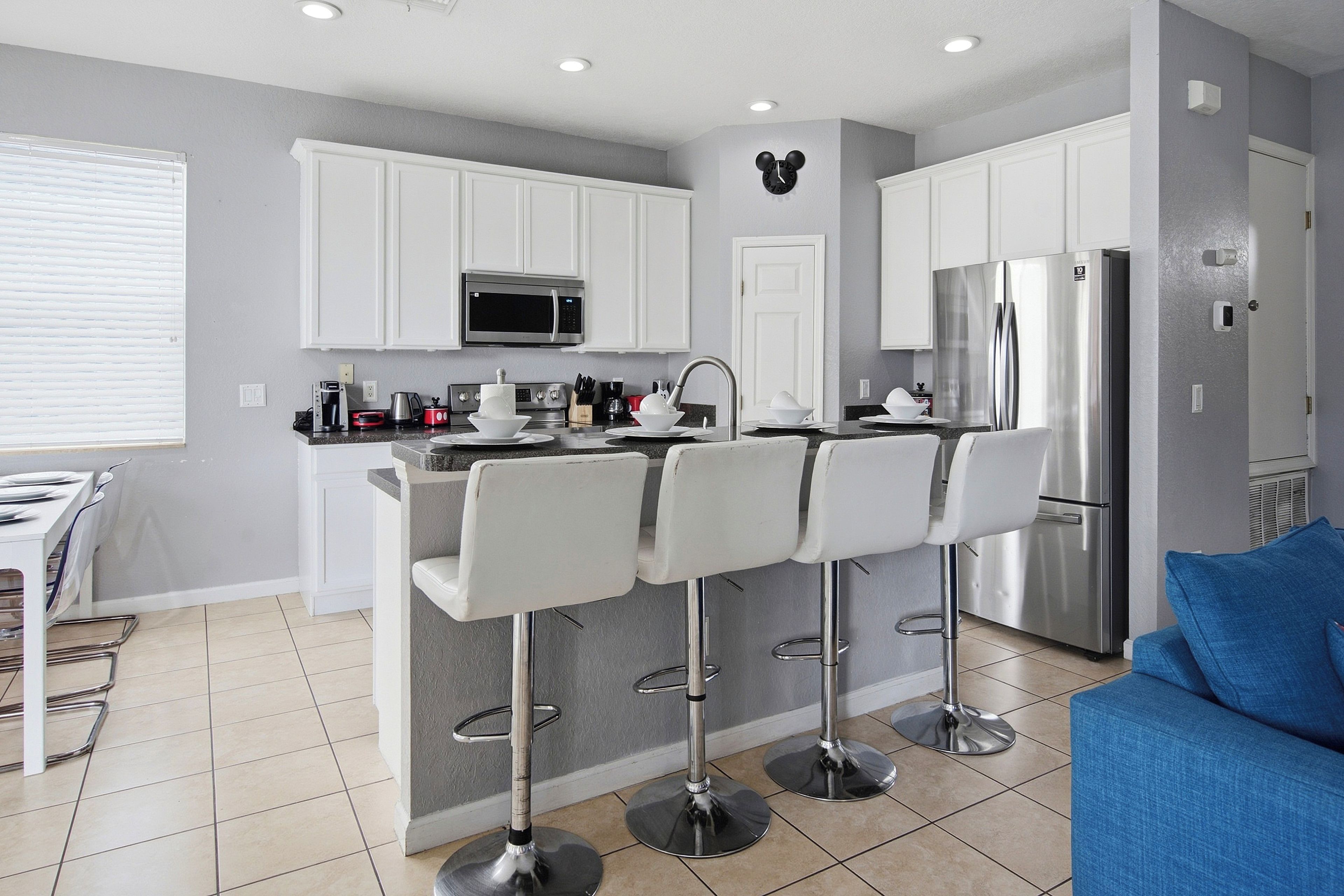 A modern kitchen with white cabinets, a stainless steel refrigerator, and a microwave. There are three white bar stools and a dining table with chairs.