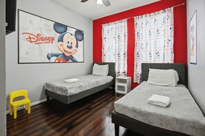 Une chambre d’hôtel avec deux lits, une affiche de Mickey Mouse, des rideaux rouges et une chaise jaune.