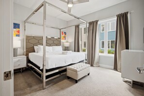 Un lit à baldaquin blanc, une table de chevet avec une lampe, un petit banc et une vue sur l’extérieur par une fenêtre avec des rideaux.