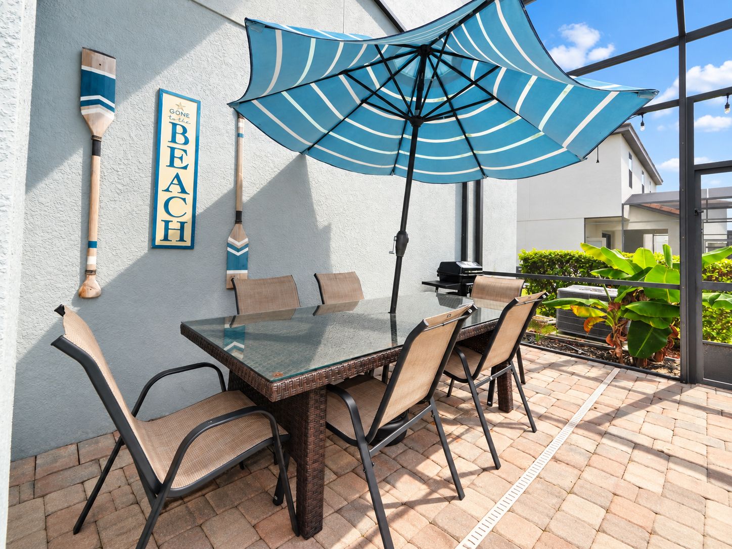 An outdoor patio with a glass table, four chairs, a blue-striped umbrella, and paddleboard decorations on the wall.