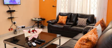 Ein Wohnzimmer mit einem schwarzen Ledersofa, einem Glastisch mit Fernbedienungen und einem an der Wand montierten Fernseher.