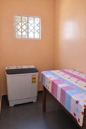 Ein kleines Zimmer mit einer Waschmaschine, einem Bett mit einer bunten Steppdecke und einem Fenster mit Gittermuster.