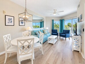 A spacious living area with a round dining table, white chairs, a blue sofa, and a ceiling fan. - Wake Up on the Gulf - Resort Life Beach Pool & Spa (Treasure Island)