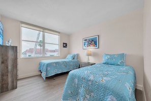 A bedroom with two beds, a window, a lamp, and a painting on the wall. - Bayfront Condo Big Views Private Beach & Pool (St. Petersburg)