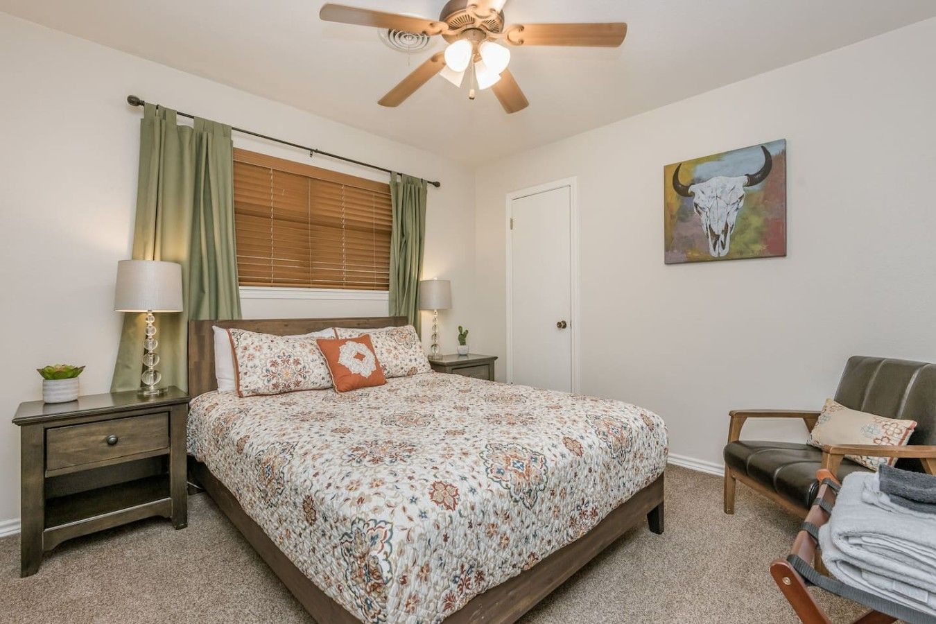 A bedroom with a bed, a nightstand, a chair, a ceiling fan, a window with curtains, and a painting on the wall.