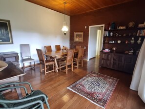 A dining area with a wooden table and chairs, a hanging light fixture, a rug, and a bookshelf with various items. - Casa Paz (Teresópolis)