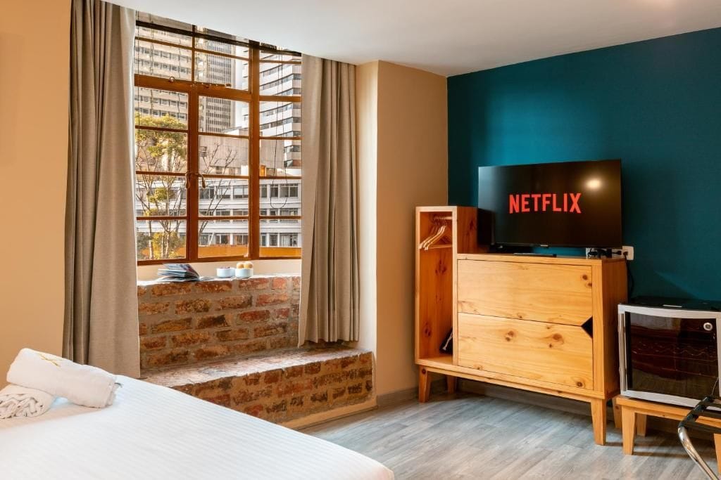 A hotel room with a large window, a wooden dresser, a television, and a small table.
