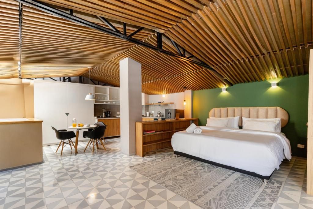 A modern hotel room with a geometric patterned floor, a wooden slatted ceiling, a bed with a white headboard, and a kitchenette area.