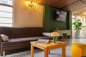 Eine moderne Lobby mit einer Couch, einem Holztisch mit Zeitschrift und Vase, einem Fernseher an der Wand und einer Pflanze.