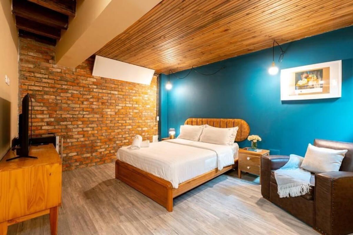Ein Schlafzimmer mit Backsteinwand, Holzboden, einem Bett mit weißer Bettwäsche, einem braunen Ledersofa und einem kleinen Tisch mit einer Lampe.
