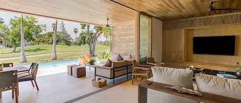 A modern living room with a large TV, comfortable seating, and a view of a swimming pool and greenery.