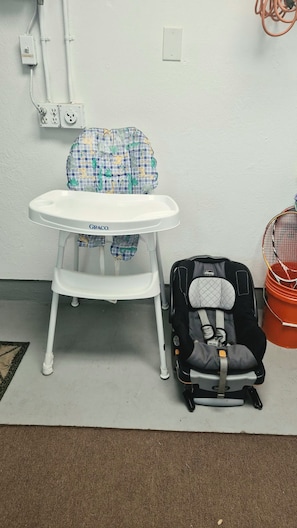 Ein weißer Hochstuhl mit kariertem Bezug, ein schwarzer Kindersitz und ein roter Korb vor einer weißen Wand mit Steckdosen.