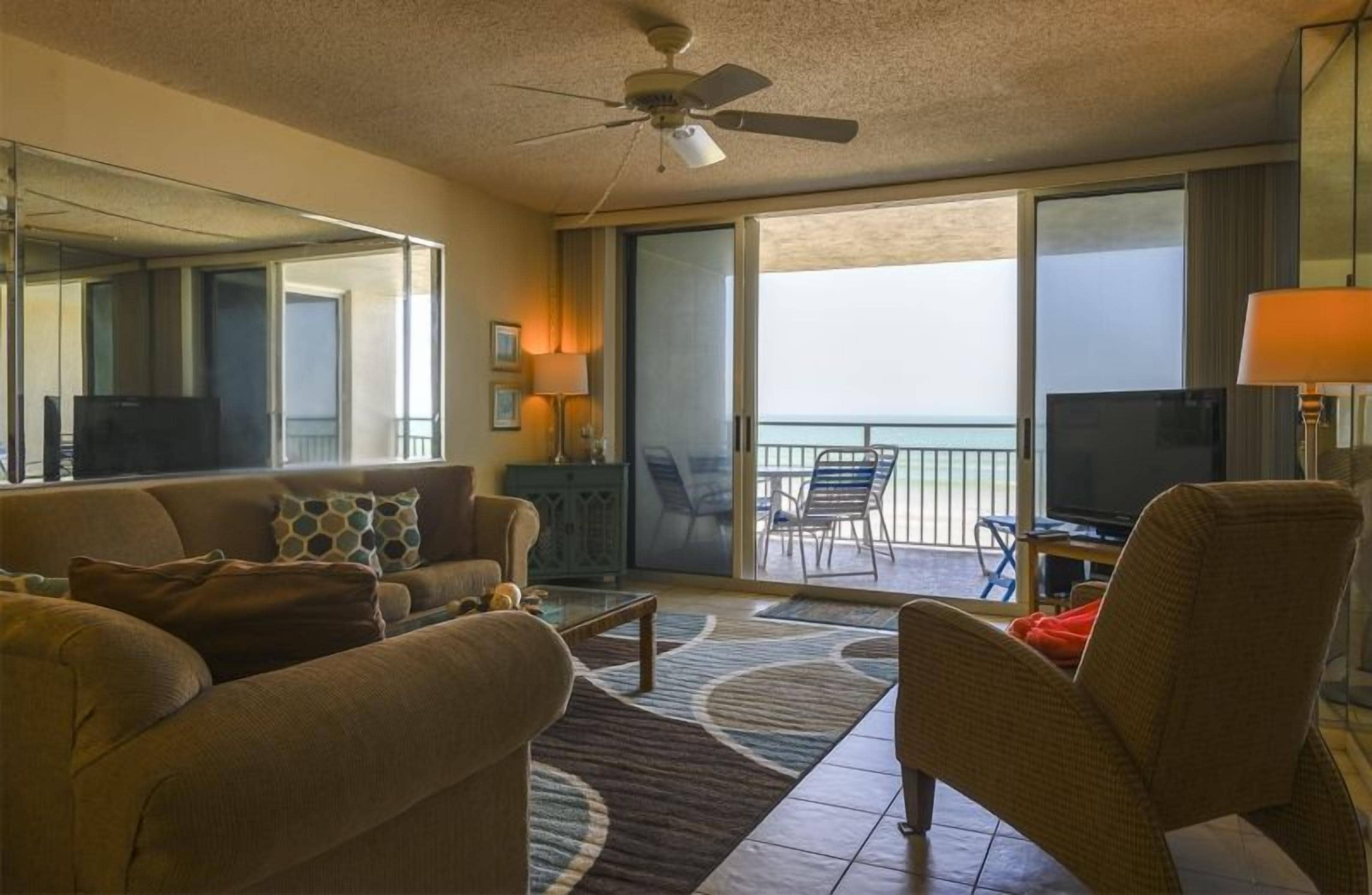 A hotel room with a sofa, armchairs, a television, a balcony with a view, and a ceiling fan.
