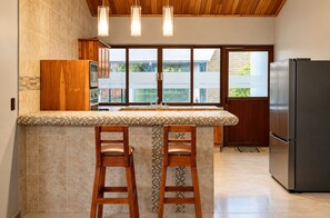 A kitchen with a large island, wooden bar stools, a refrigerator, and a view to the outside.