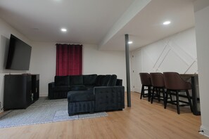 Ein modernes Wohnzimmer mit einer schwarzen Couch, einem kleinen Ottomanen, einem Flachbildfernseher und einem Essbereich mit drei Stühlen.