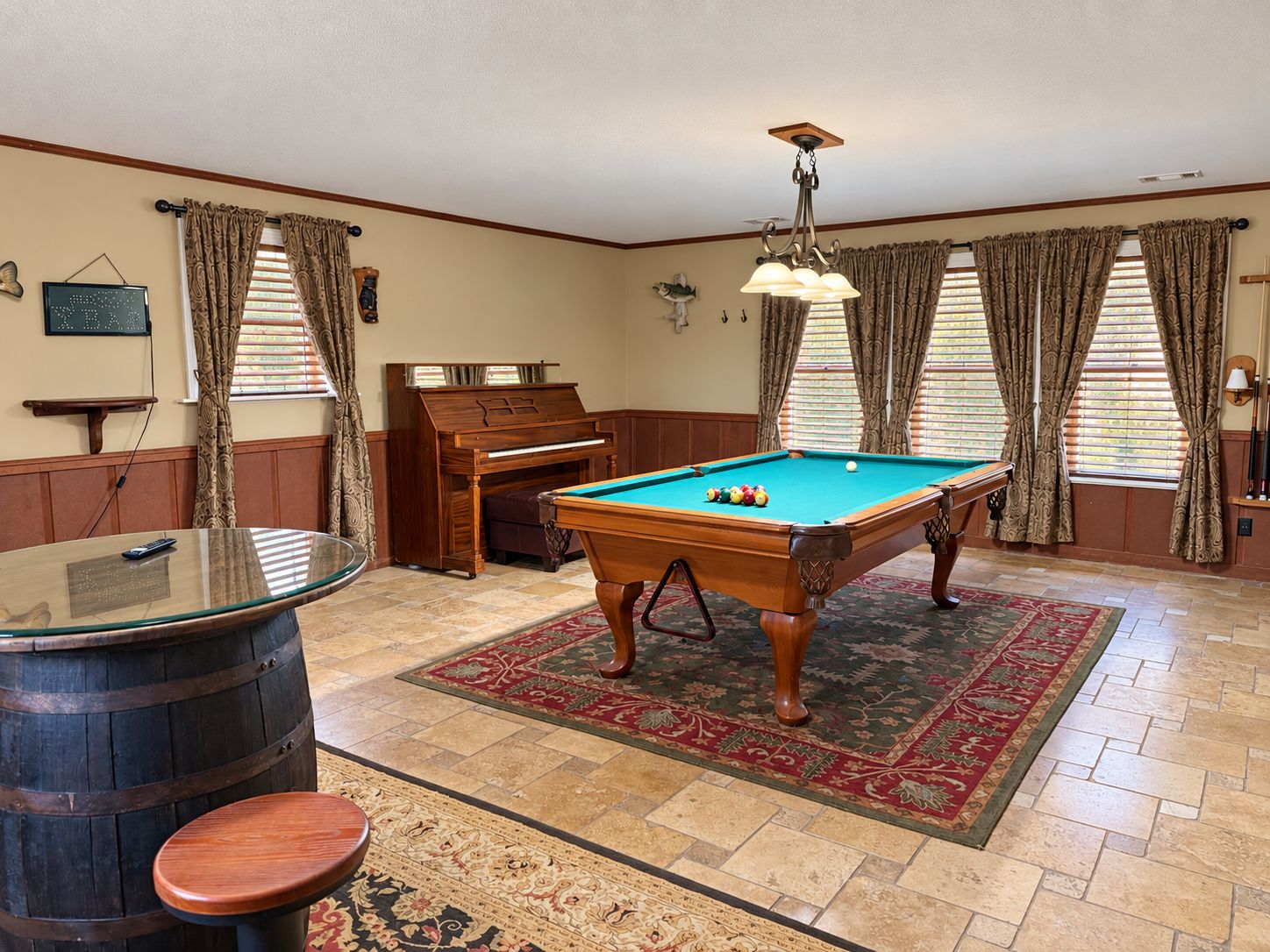 A pool table with a billiard cue, a piano, and a bar stool in a room with patterned curtains and a red carpet.