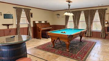 A pool table with a billiard cue, a piano, and a bar stool in a room with patterned curtains and a red carpet.