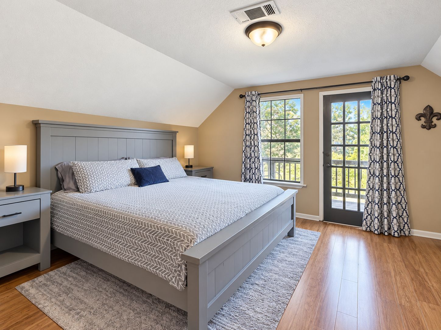 A bedroom with a bed, nightstands, a window with curtains, and a ceiling vent.