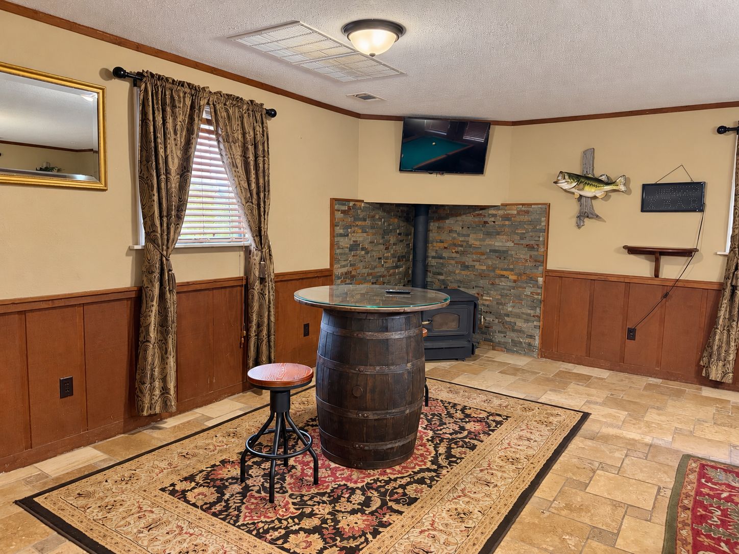 A room with a stone fireplace, a round wooden barrel, a small stool, a television mounted on the wall, and a patterned rug on the floor.